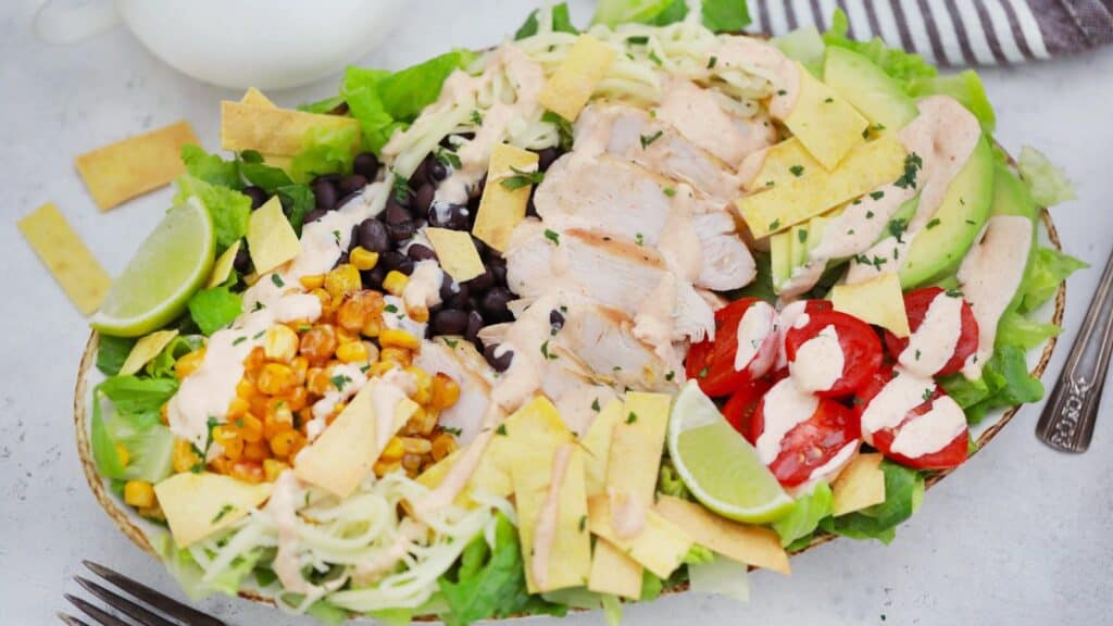 A vibrant chicken salad with avocado, black beans, corn, cherry tomatoes, and lime slices arranged neatly on a white plate.