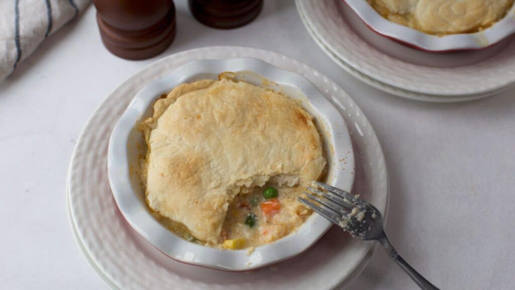 A seafood pot pie with a golden crust served in a white bowl, with a fork taking a bite out, revealing the creamy filling with vegetables.
