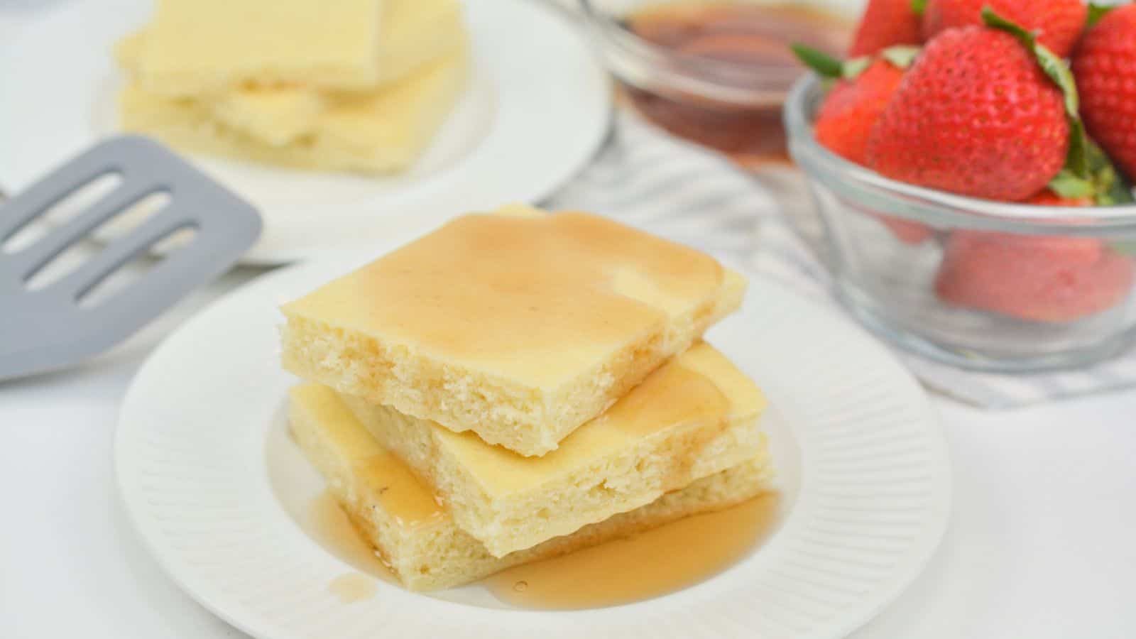 A plate of square pancakes drizzled with syrup, next to strawberries and a cup of maple syrup on a white tablecloth.