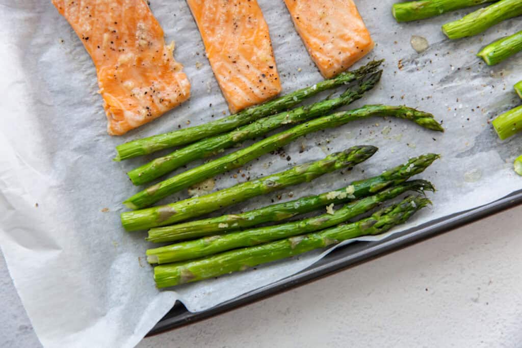 Four grilled salmon fillets and a row of asparagus spears on parchment paper on a baking tray.