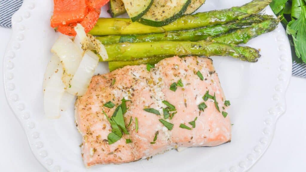 Baked salmon fillet with asparagus, zucchini, and bell peppers on a white plate, garnished with herbs.