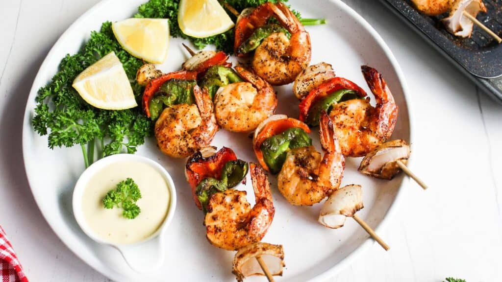 Grilled shrimp skewers with red and green bell peppers, served with lemon wedges and dipping sauce on a white plate.
