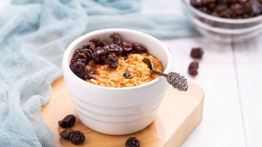 A bowl of baked oatmeal topped with raisins on a wooden table, accompanied by a blue cloth and spoon.