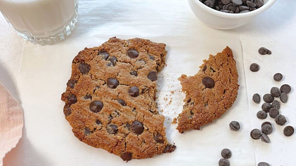 A large chocolate chip cookie with a piece broken off, next to a glass of milk and a bowl of chocolate chips on a white surface.