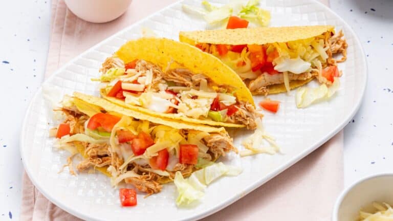 Three chicken tacos with shredded cheese, lettuce, and diced tomatoes on a white plate.