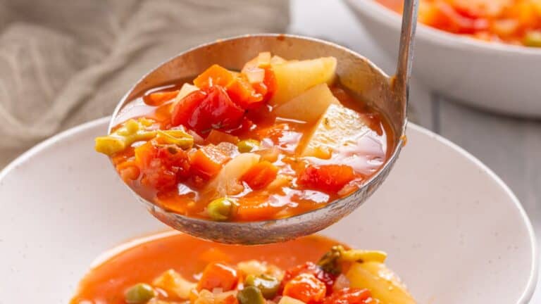 A ladle serving vegetable soup with tomatoes, potatoes, and green peas into a white bowl, with a blurred bowl in the background.