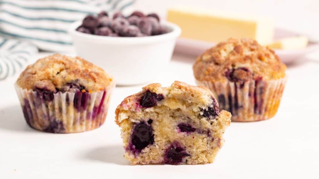 Two blueberry muffins, one cut in half, on a plate with fresh blueberries and slices of butter in the background.