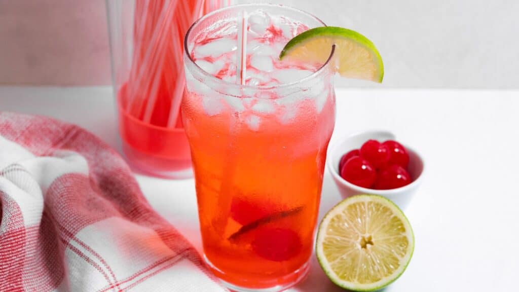 A glass of red iced tea with a lime wedge, next to a bowl of cherries and a pitcher filled with homemade versions of the same beverage.
