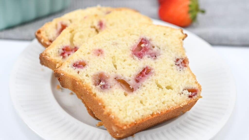 Three slices of strawberry pound cake on a white plate, displaying visible chunks of strawberries, with a blurred background.