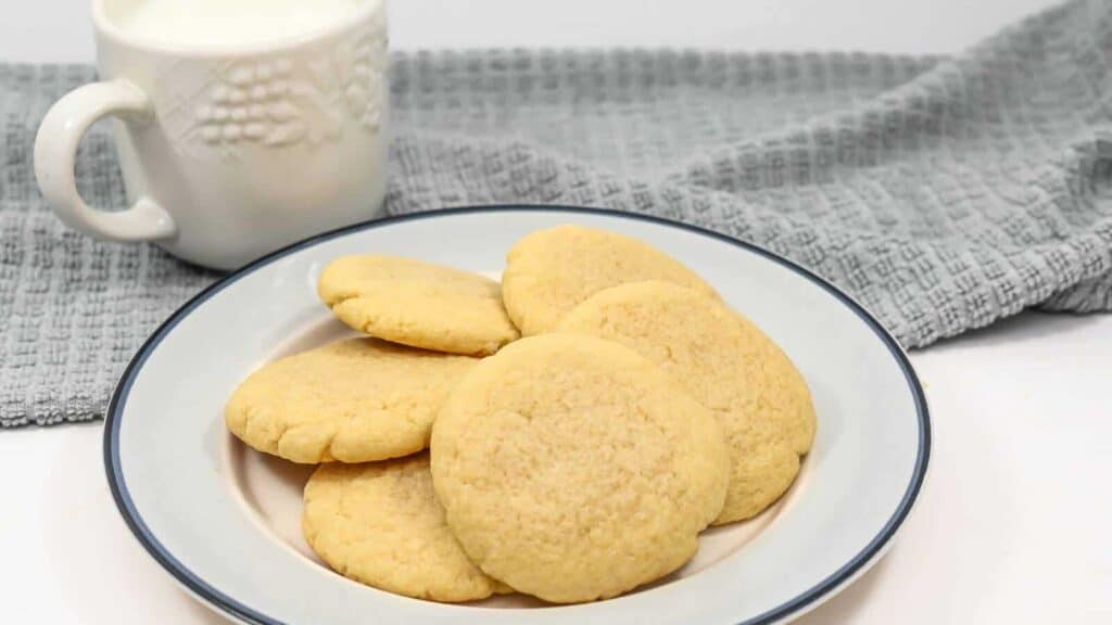 A plate of sugar cookies beside a white mug and a grey cloth on a white background.