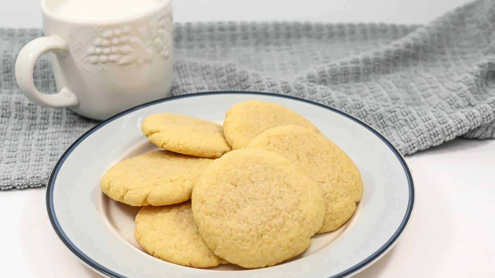 A plate of sugar cookies with a mug of milk and a gray cloth in the background.