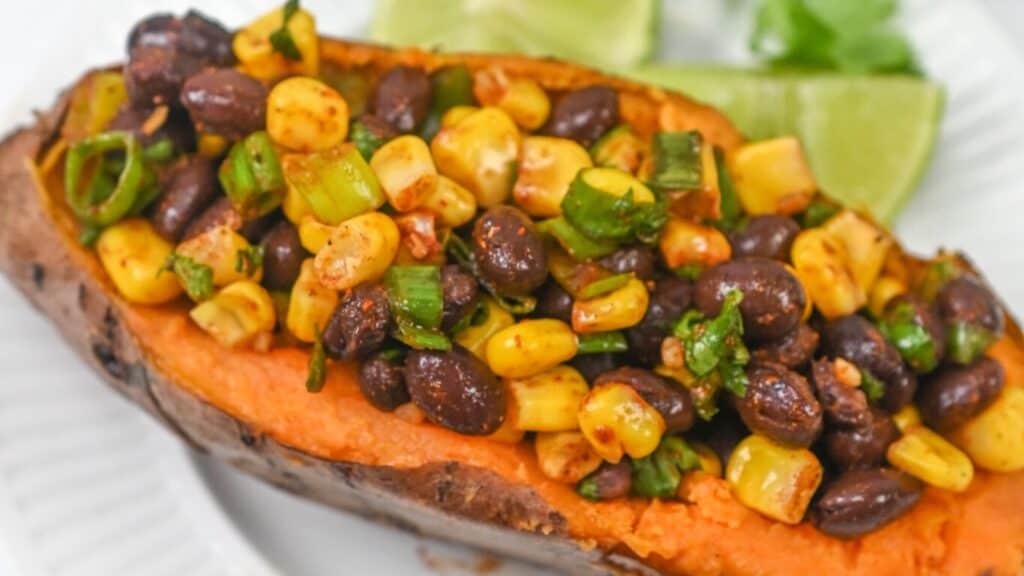 Stuffed sweet potato with black beans, corn, and green onions garnished with lime wedges on a white plate.
