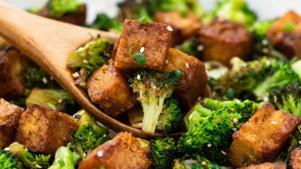 Roasted broccoli and tofu cubes garnished with sesame seeds and green onions, served with a wooden spoon.
