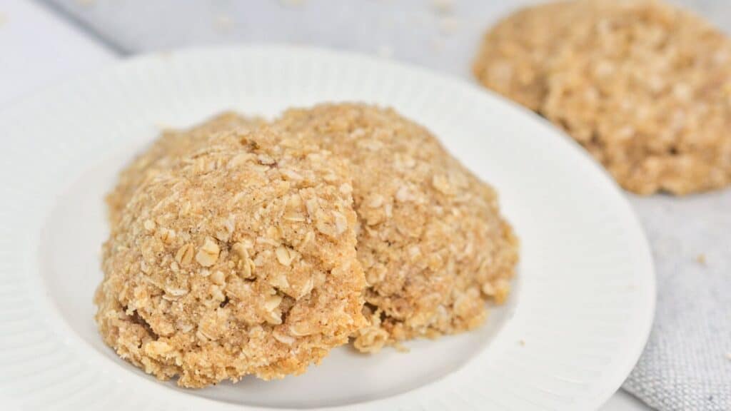 Two oatmeal cookies on a white plate, with a crumbly texture and visible oats, displayed on a light-colored tablecloth.