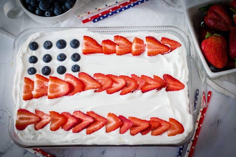 A rectangular cake decorated to resemble the American flag, using blueberries for stars and sliced strawberries for stripes, placed on a white cloth with bowls of strawberries and blueberries nearby.