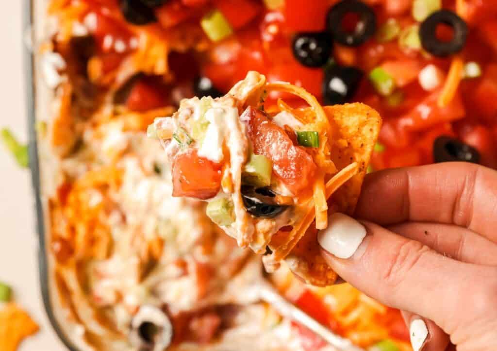 A hand holding a chip loaded with toppings, including diced tomatoes, black olives, green onions, shredded cheese, and a creamy sauce, lifted from a tray filled with similarly topped nachos.