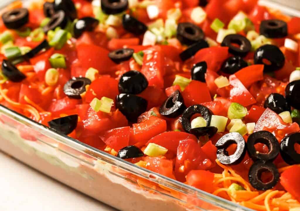 A close-up of a seven-layer dip topped with black olives, diced tomatoes, and sliced green onions in a glass dish.