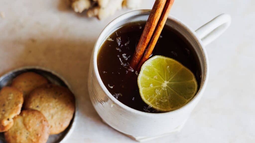 A hot toddy in a white mug with a cinnamon stick and lime slice, accompanied by a plate of cookies.