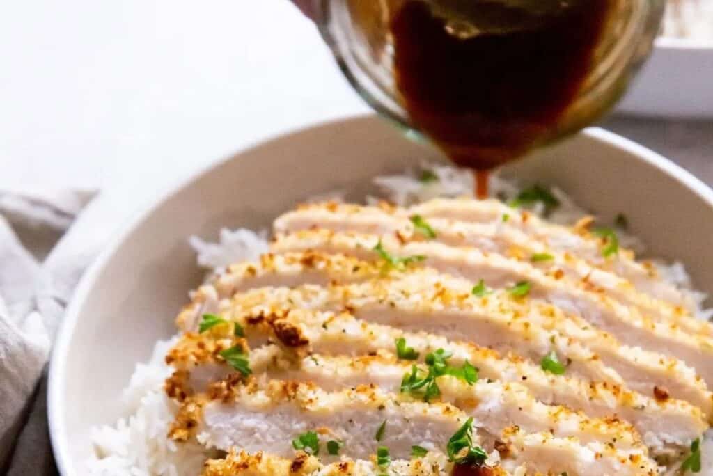 Gravy being poured over sliced chicken breast on a bed of white rice, garnished with herbs.