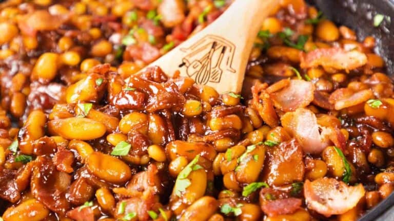 A close-up of baked beans with bacon and chopped parsley in a skillet, a wooden spoon resting inside.