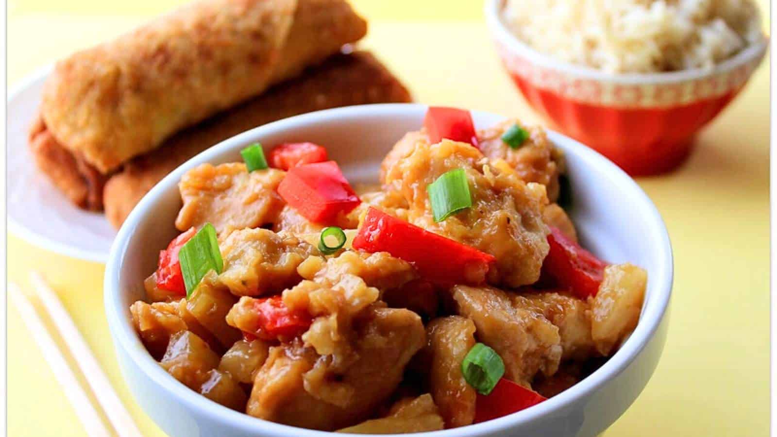 A bowl of sweet and sour chicken with red bell peppers, garnished with green onions, accompanied by a spring roll and a small bowl of rice.