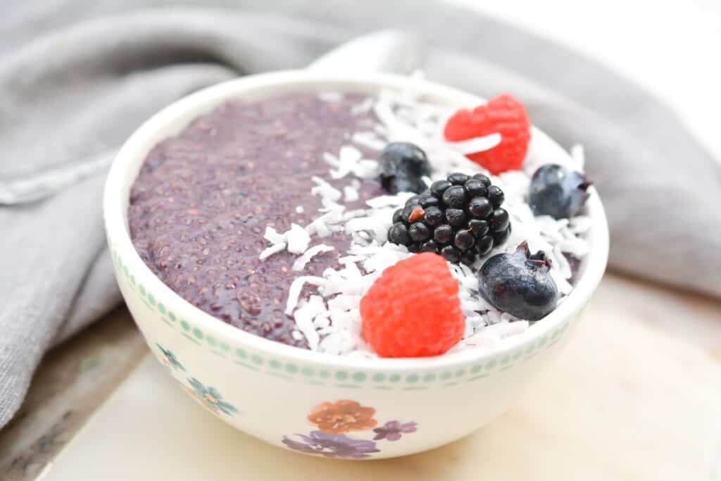 A bowl of chia pudding topped with raspberries, blackberries, blueberries, and shredded coconut, with a gray cloth in the background.