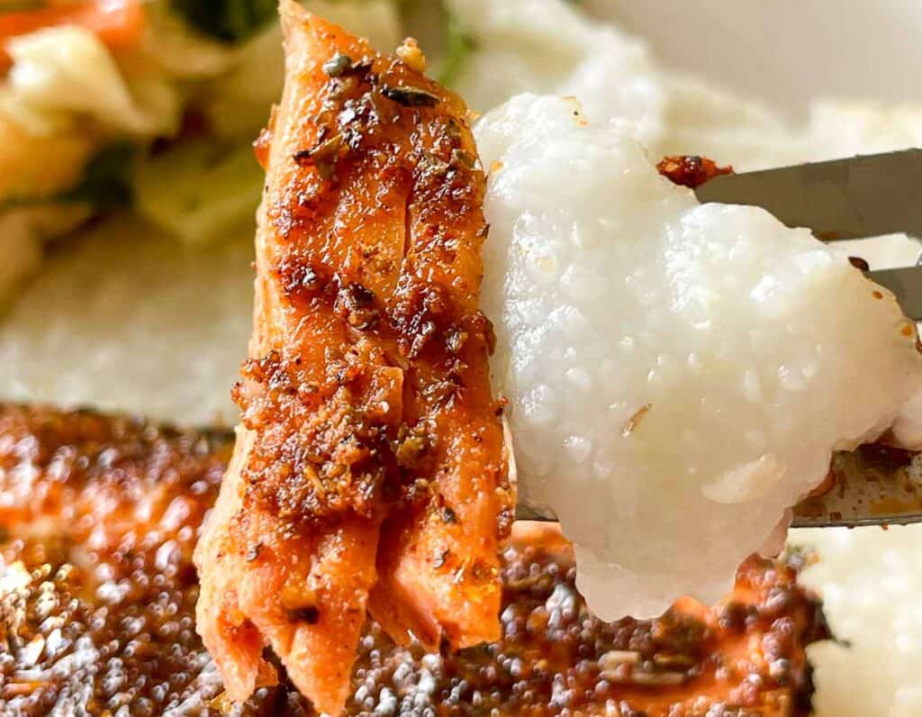 A fork holds a piece of seasoned, cooked fish and a portion of white grits, with a side of vegetables visible in the background.