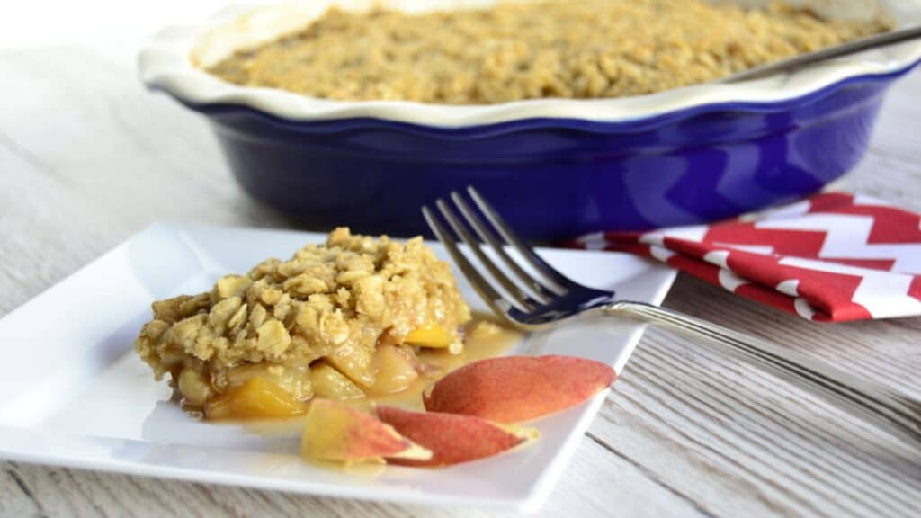 A slice of peach crumble on a square white plate, with a fork, peach slices, and a blue baking dish filled with more crumble in the background.