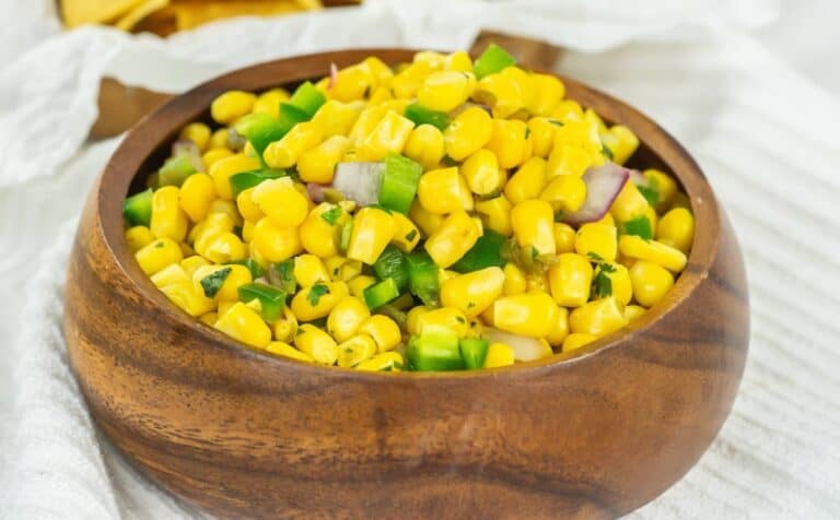 A wooden bowl filled with corn salad, containing diced green bell peppers, onions, and herbs, served on a white cloth.