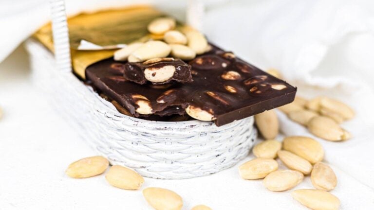 A bar of dark chocolate with nuts inside a small white woven basket, surrounded by scattered almonds on a white surface.