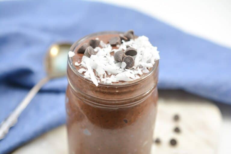 A glass jar filled with chocolate chia seed pudding topped with shredded coconut and chocolate chips, placed on a table with a spoon and a blue cloth in the background.