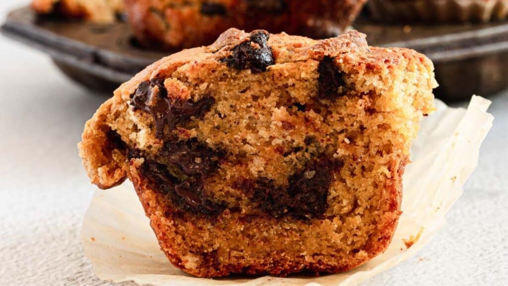 Close-up of a chocolate chip muffin with a bite taken out, revealing the inside texture and chocolate chunks. The muffin rests on a white wrapper with a muffin tray in the background.