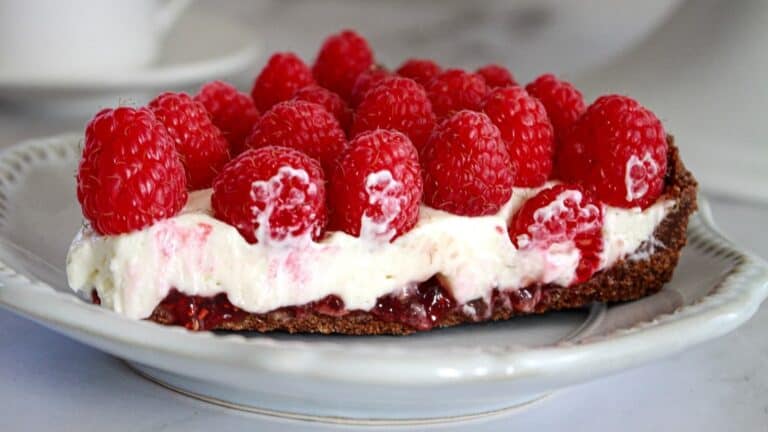 A plate with a slice of tart topped with a thick layer of cream cheese and raspberry jam, garnished with fresh raspberries.