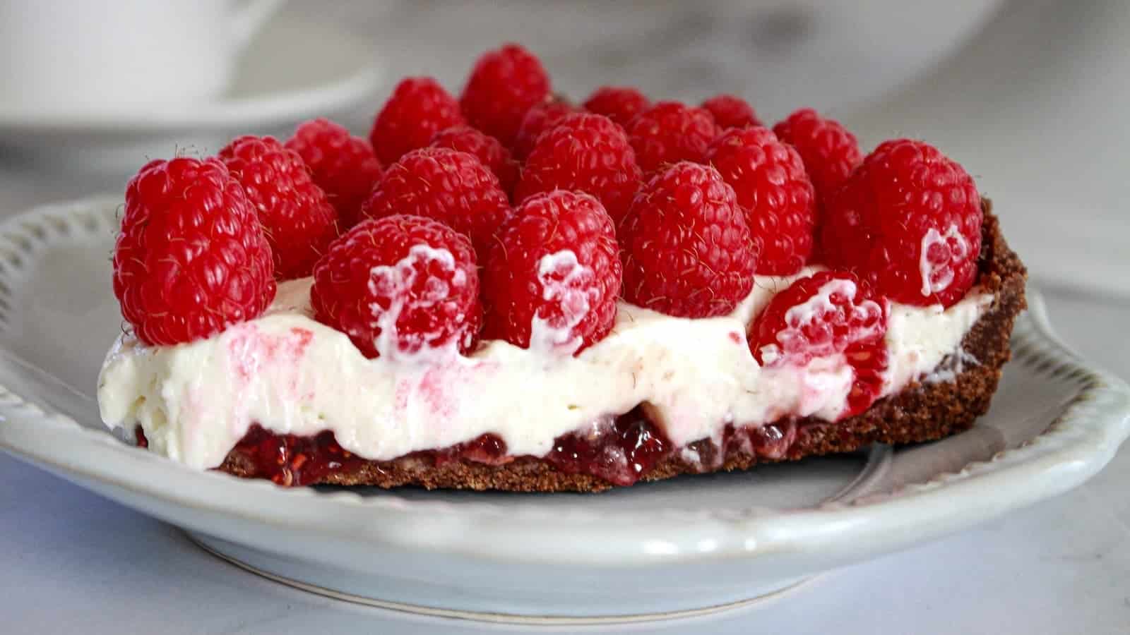 A slice of tart topped with a layer of jam, cream, and whole raspberries on a ceramic plate.