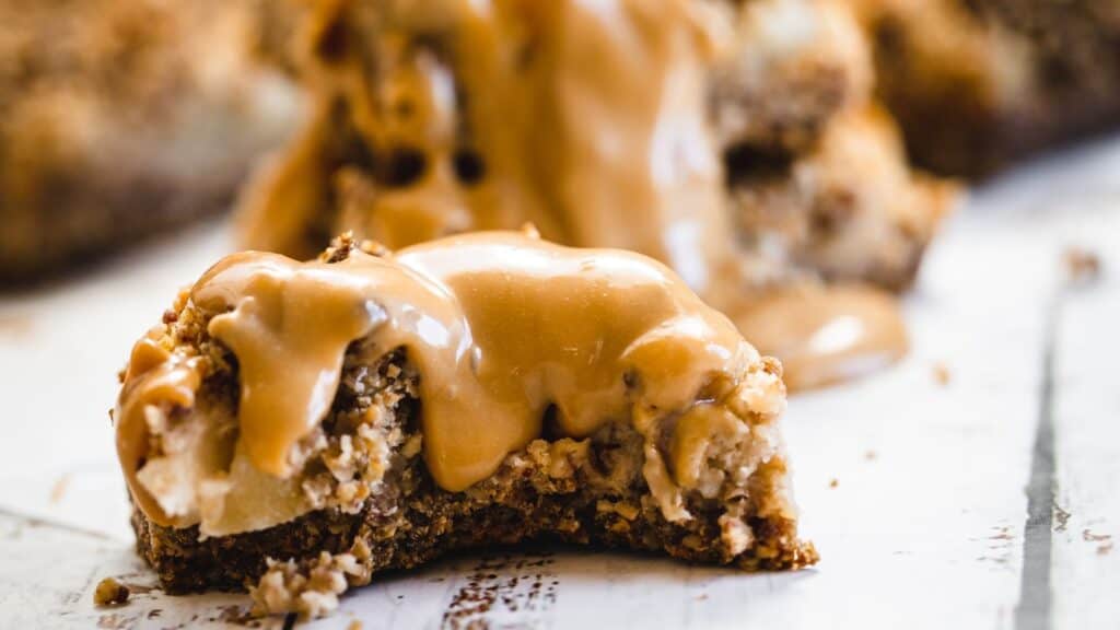 A close-up image of a cookie drizzled with caramel, with visible chunks of nuts, set on a light wooden surface.