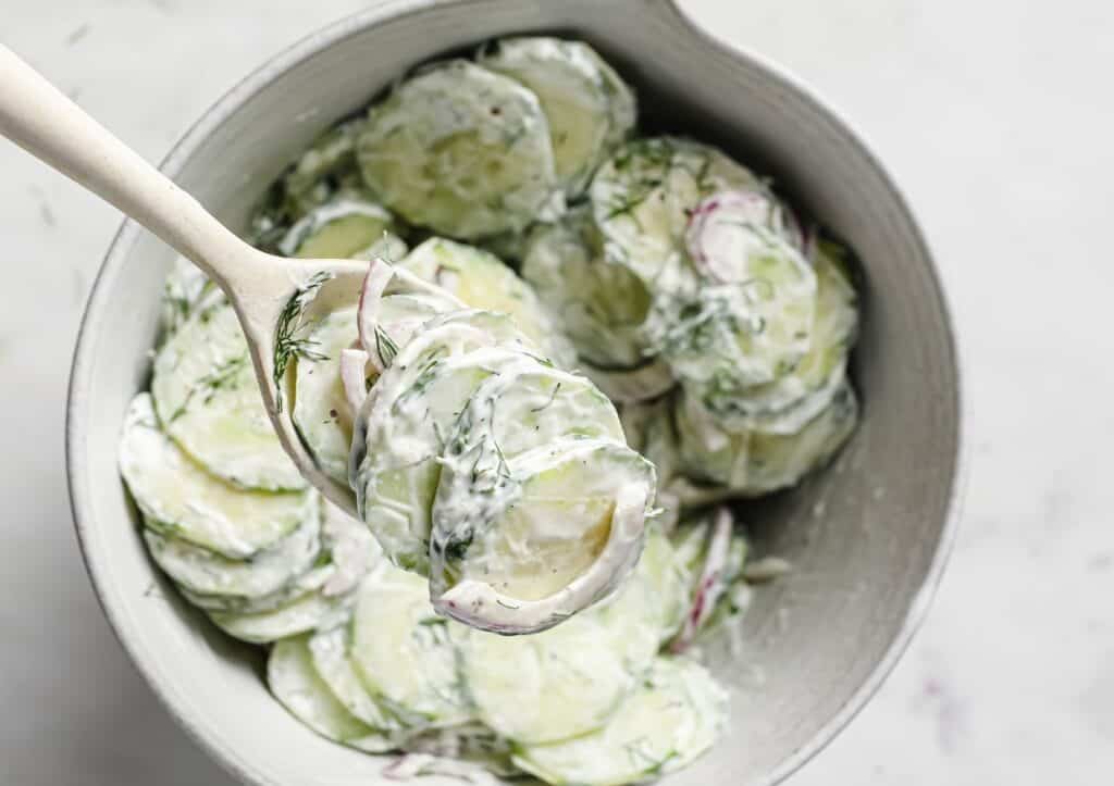 A bowl of creamy cucumber salad with fresh dill and red onion, being lifted with a wooden spoon.