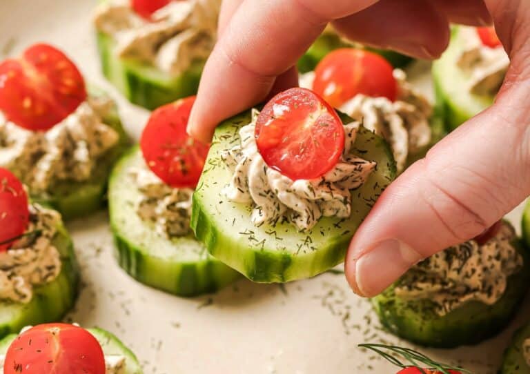 A hand holds a cucumber slice topped with cream cheese, dill, and half a cherry tomato. Other similar cucumber appetizers are arranged on a plate.