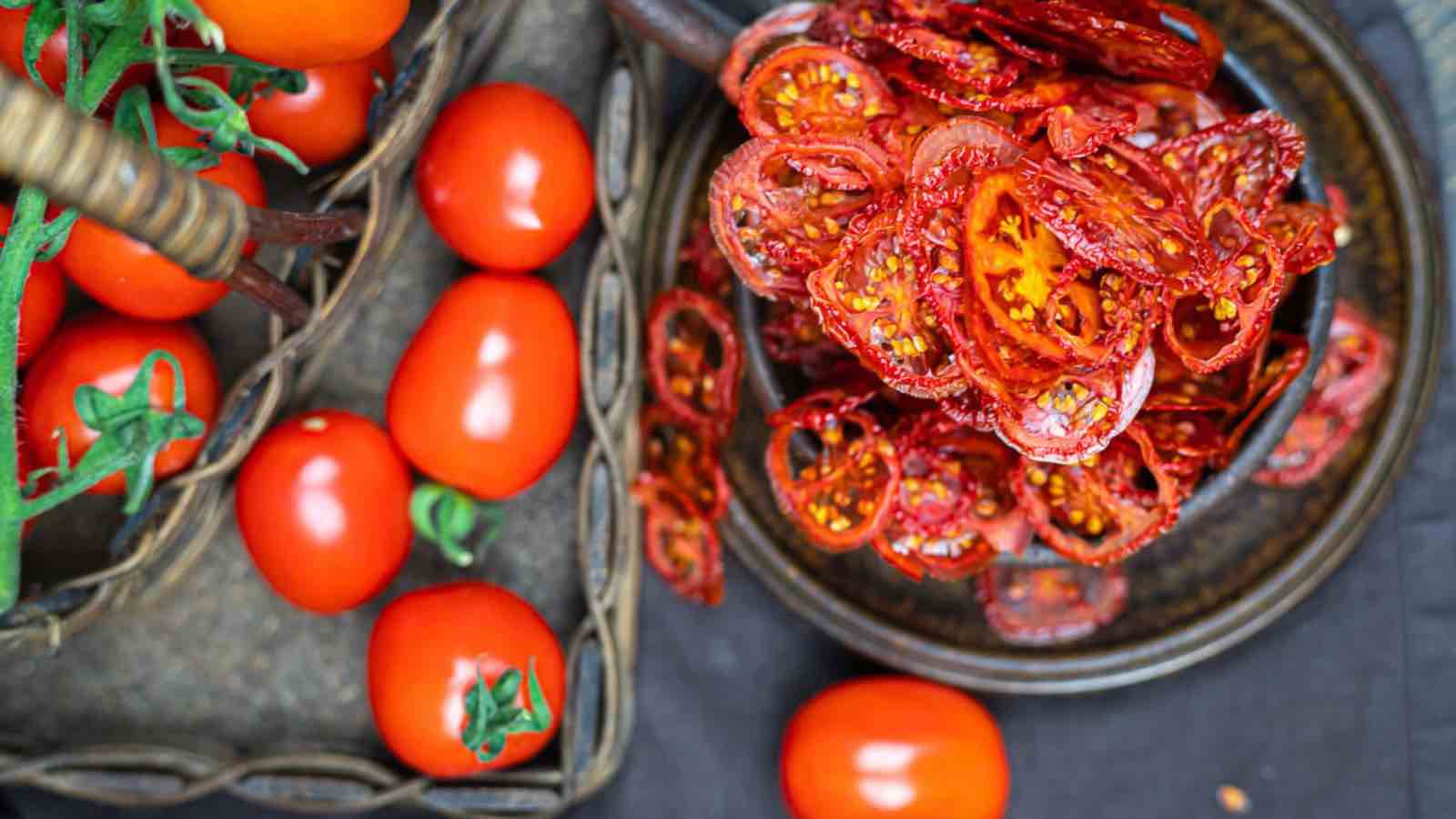A basket of fresh whole tomatoes next to a plate of sliced, sun-dried tomatoes on a dark textured surface.