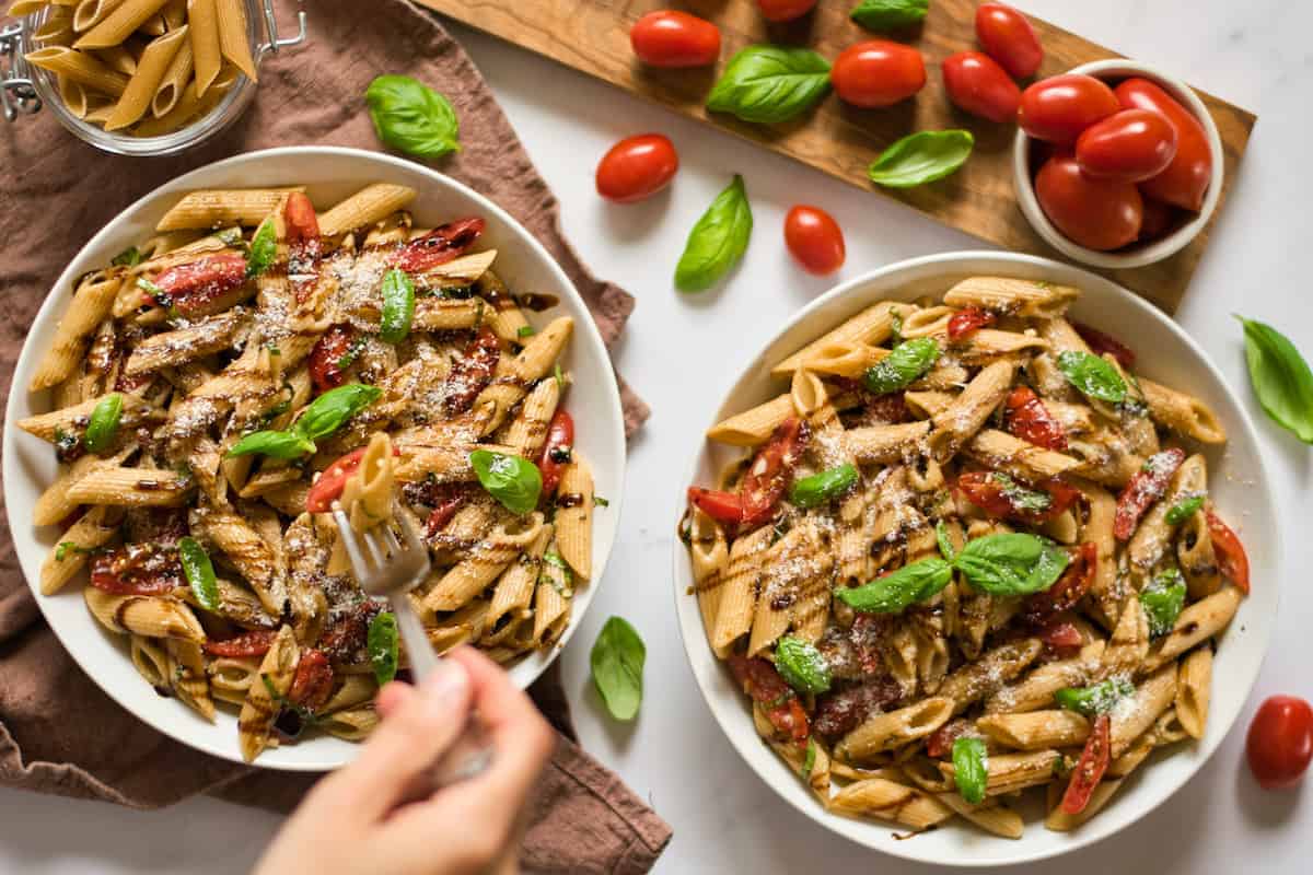 Two bowls of penne pasta with cherry tomatoes, basil, and grated cheese, drizzled with balsamic reduction. A hand holding a fork is reaching into one of the bowls. Fresh tomatoes and basil are nearby.