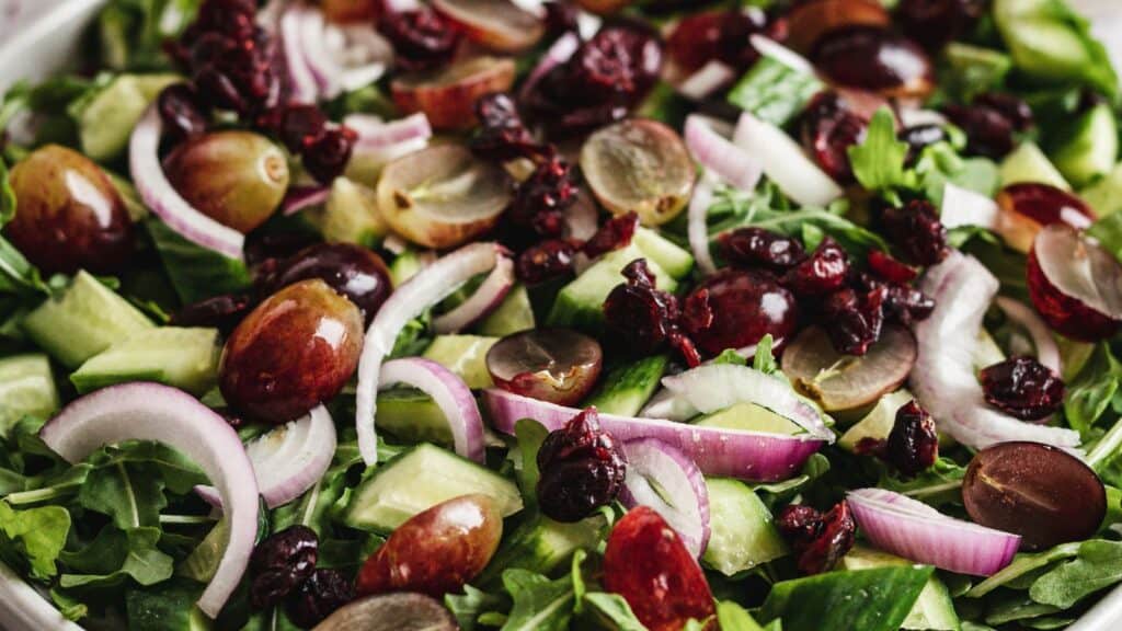 Close-up of a fresh salad featuring mixed greens, sliced red grapes, cucumbers, red onion, and dried cranberries.