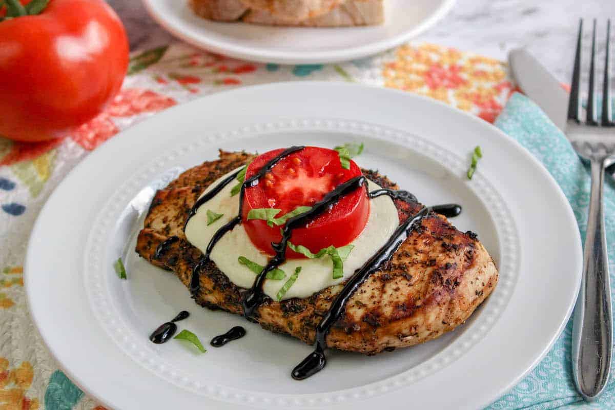 Grilled chicken breast topped with a slice of mozzarella, a tomato slice, balsamic glaze, and chopped basil, served on a white plate with a fork and knife nearby.