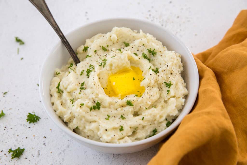 A bowl of mashed potatoes garnished with parsley and a butter pat melting in the center, accompanied by a spoon and an orange napkin on a white surface.