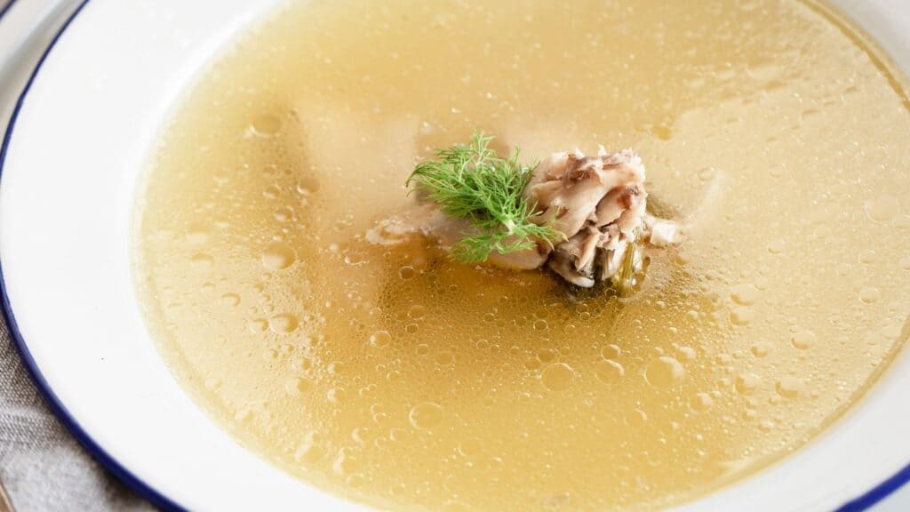 A bowl of clear soup with chicken pieces and dill, served in a white bowl with a blue rim.