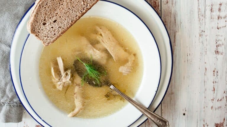 A bowl of clear broth with chicken, herbs, and a slice of bread on a rustic wooden table.