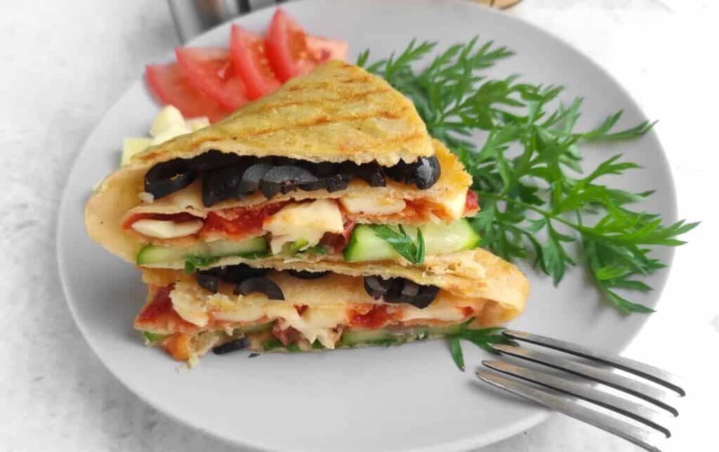 A quesadilla filled with cheese, tomatoes, cucumbers, and olives on a plate with fresh parsley and diverse treats of tomato slices on the side.