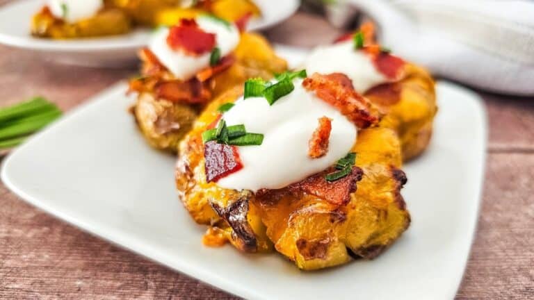 A close-up of crispy smashed potatoes topped with sour cream, chopped chives, and crispy bacon pieces, served on a white plate.