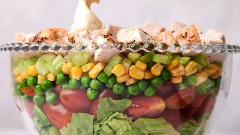 A glass bowl filled with layered salad, including chopped lettuce, cherry tomatoes, peas, corn, diced celery, and grilled chicken pieces, topped with a dollop of mayonnaise.