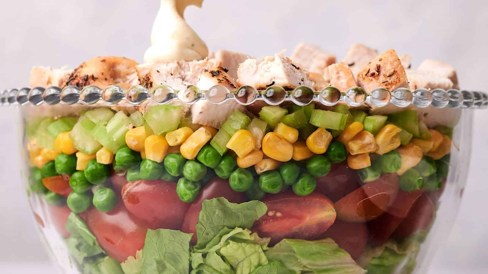 A clear glass bowl layered with lettuce, cherry tomatoes, green peas, corn, diced celery, and topped with chunks of grilled chicken, garnished with a dollop of mayonnaise.