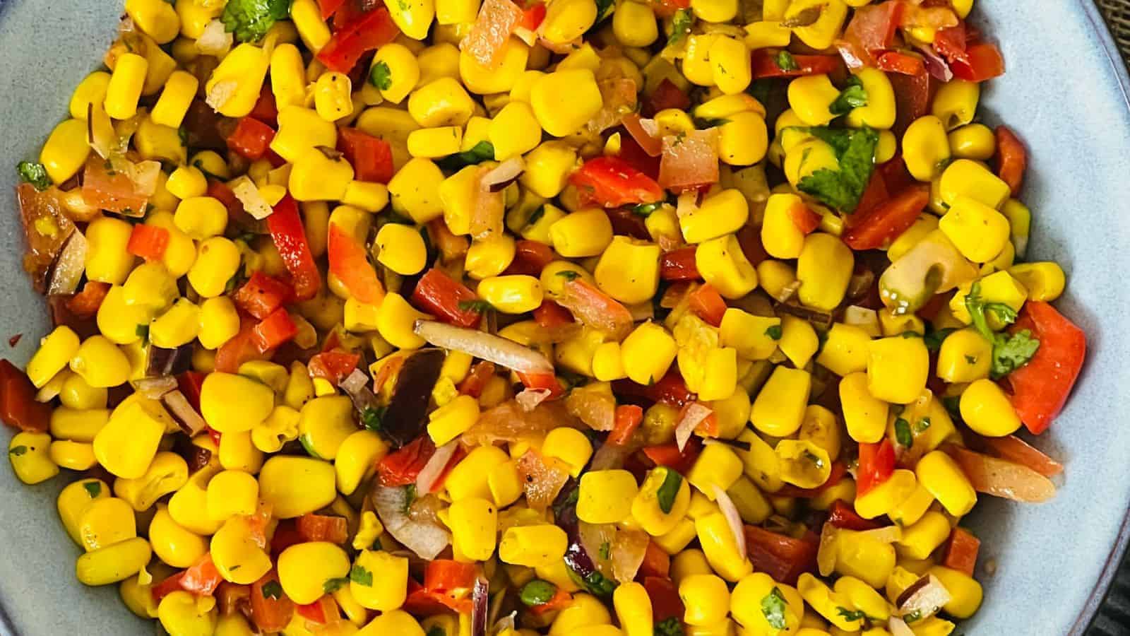 A close-up of a bowl filled with corn salad, featuring yellow corn kernels mixed with diced red bell peppers, onions, and herbs.