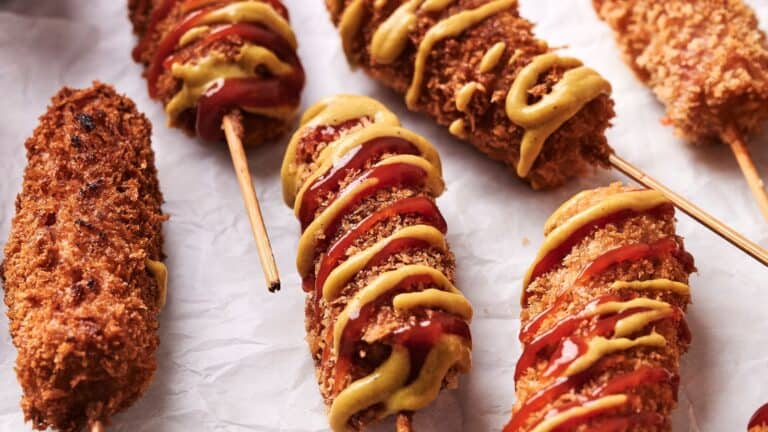 Several breaded and deep-fried corn dogs are shown on a sheet of parchment paper, topped with mustard and ketchup.